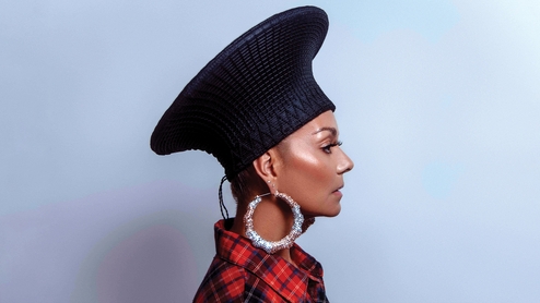 Photo of Anané. She's posing in profile against a grey background. She's wearing a red flannel shirt, large hoop earrings and a dark hat with a wide rim