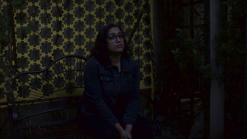Photo of Krithi sitting on a black bench at night in front of a yellow and brown patterned curtain