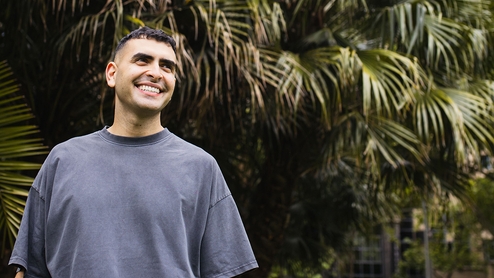 Sammy Virji standing in a washed blue t-shirt in front of plants. He is smiling and looking away from the camera.