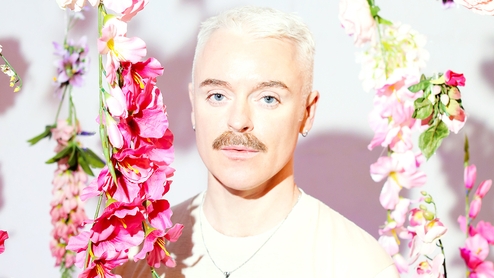A brightly lit photo of Cormac in a white t-shirt next to a variety of pink hanging flowers
