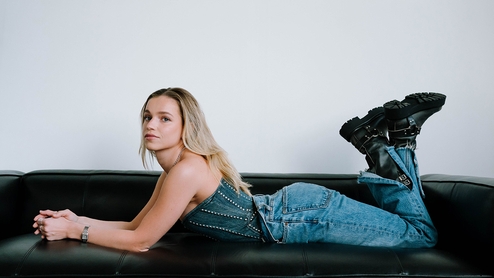 Press shot of Jackie Hollander lying on her front on a black couch wearing denim jeans, denim top and black boots