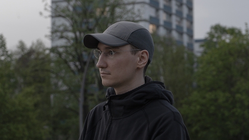 Photo of buttechno standing outside on an overcast day near an apartment block and trees. He's wearing a grey baseball cap and glasses