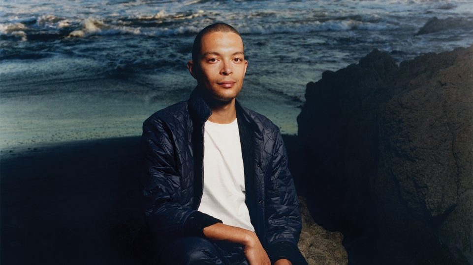 Batu press shot against the sea in a blue quilted jacket and white t-shirt