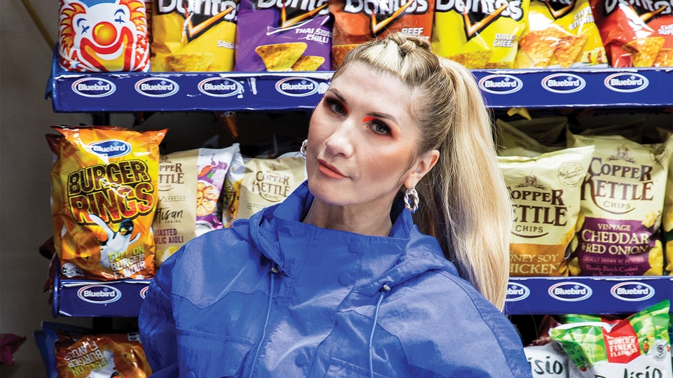 Tali in a blue coat in front of a row of crisp packets