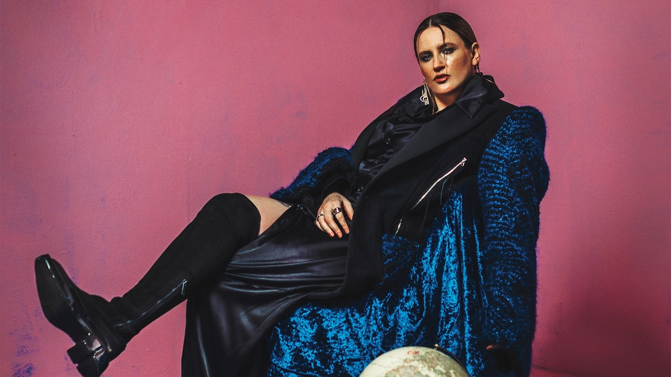 Saoirse reclining on a stool in a long blue velvet coat and knee high black boot
