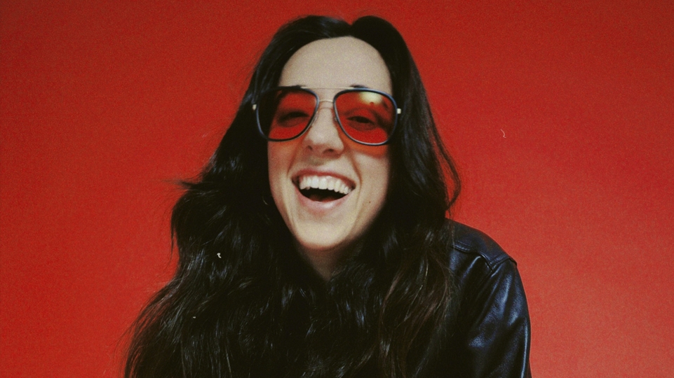 Chloé Caillet smiling in red sunglasses against a red backdrop