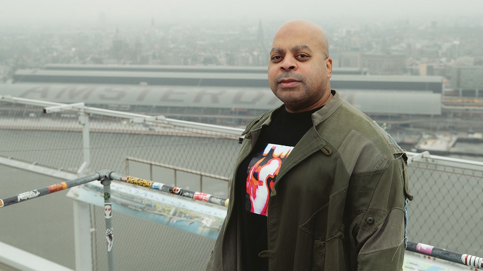 Photo of DJ Bone standing on a tall roof overlooking Amsterdam train station in a green jacket