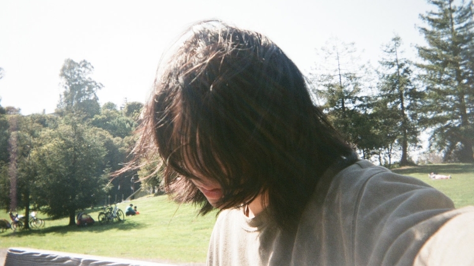 Photo of Tomu DJ gazing downwards with long hair sweeped in front of her face in a green park space