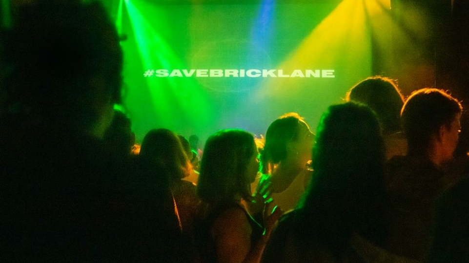 Crowd shot taken inside a club night with the words #SAVEBRICKLANE projected on stage