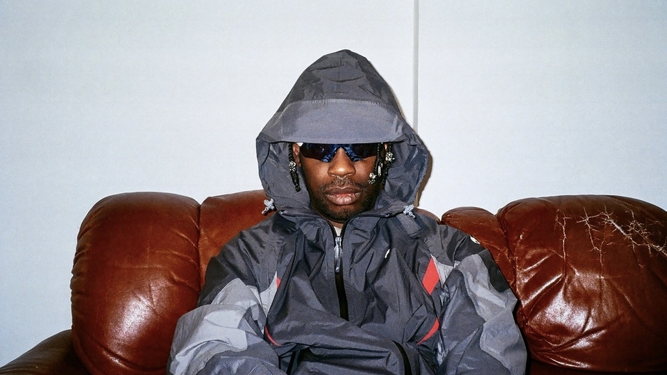 Photo of GAIKA wearing a grey parka and sunglasses while sitting on a sofa