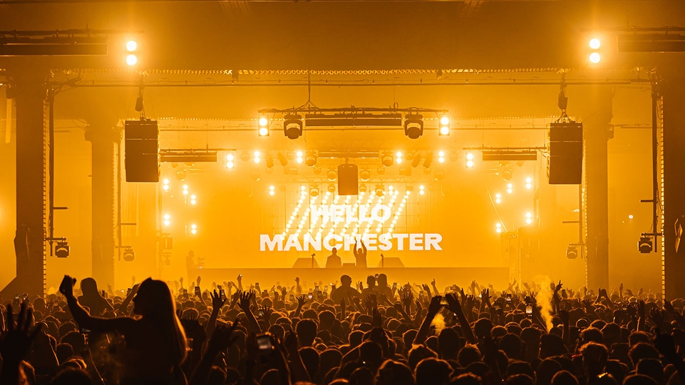 Photo of gold lights and a crowd inside a The Warehouse Project event. The words "hello Manchester" are projected on the stage