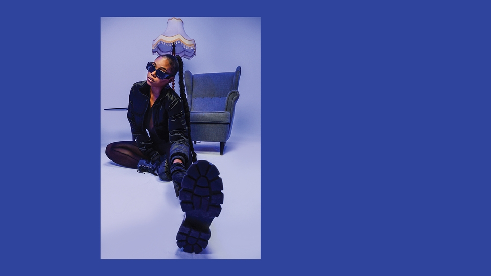 Melanin Tee wearing a black leather coat and boots sitting on the floor infront of a chair. Her hair is braided, she wears sunglasses, and one leg is stretched toward the camera