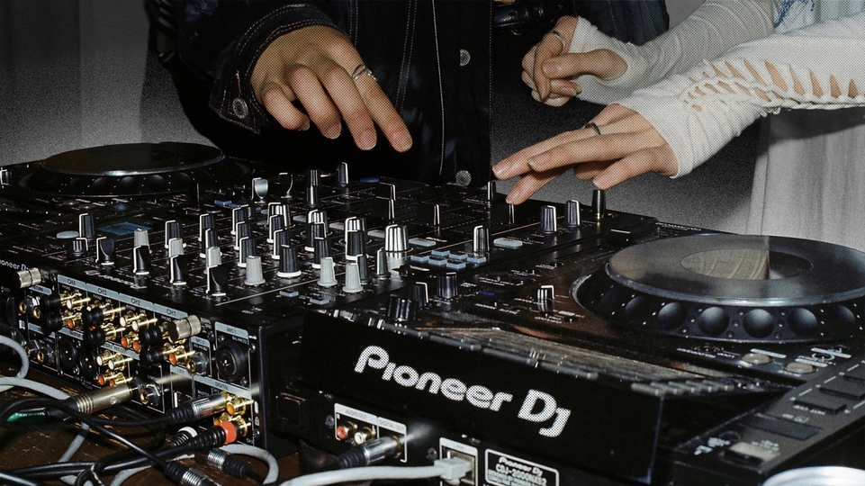 Close up image of two CDJs and a mixer being used by two people, whose hands are visible
