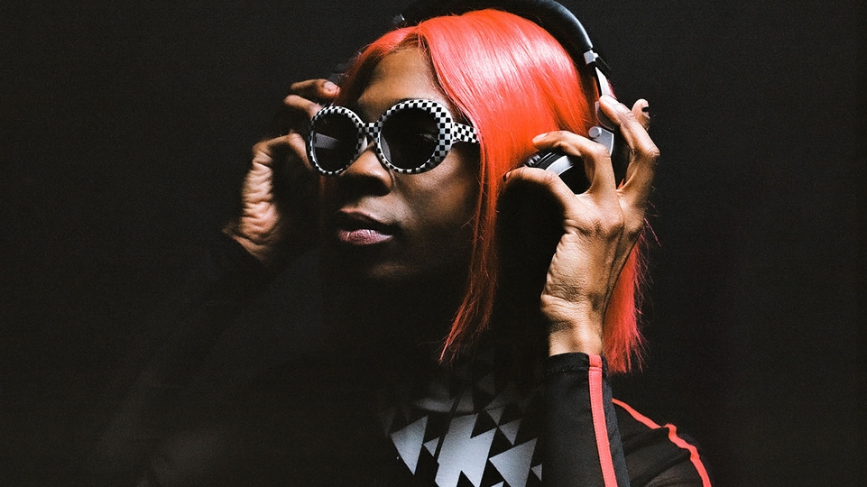 Photo of quest?onmarq posing against a black background. Her hair is long and dyed red and she's holding a pair of headphones over her ears. She has a pair of black and white sunglasses on