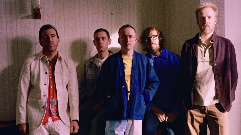 Photo of the five members of Hot Chip standing in front of a wall in a house with a pink hue