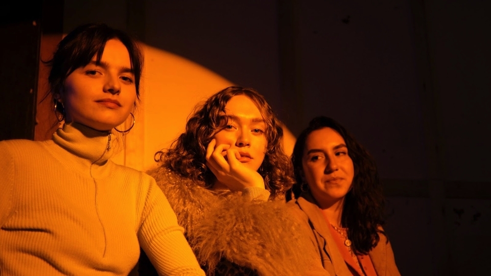 LOTTIE PENDLEBURY, LENNY WATSON, SOPHIE FARRELL of SISTER MIDNIGHT sat against a wall with evening sun shining on them through a window