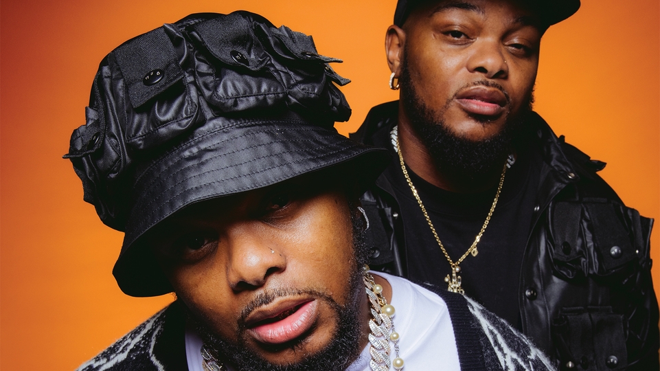 Photo of Bandile and Banele posing wearing black hats in front of a bright orange background