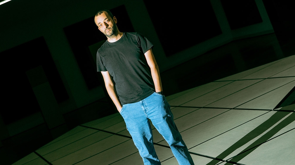ABADIR standing in a large room with white tiles. The room is dark and he is under a large spotlight, wearing blue jeans and a dark grey t-shirt.
