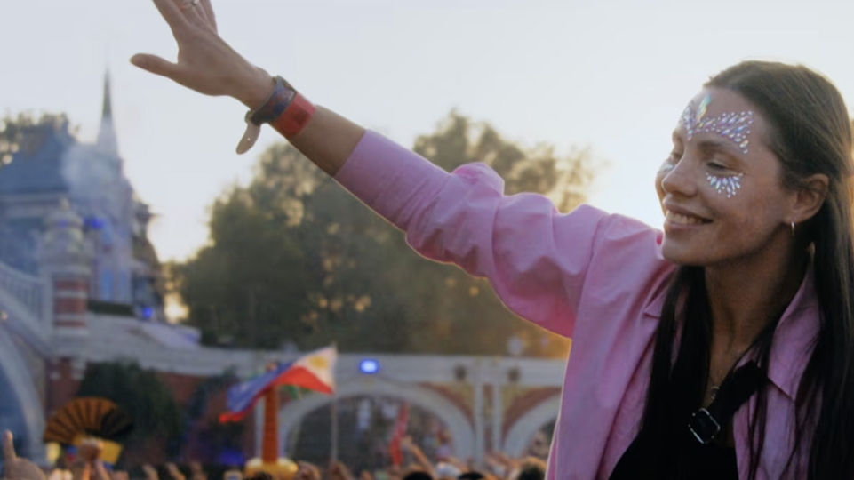 Photo of a woman in a pink shirt and long brown hair with glittery make up dancing with her arm outstretched in the Tomorrowland crowd