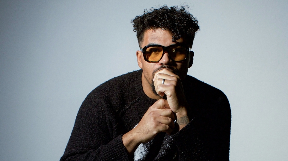 Photo of DJ Cuddles wearing orange sunglasses and a black sweatshirt in front of a light blue background