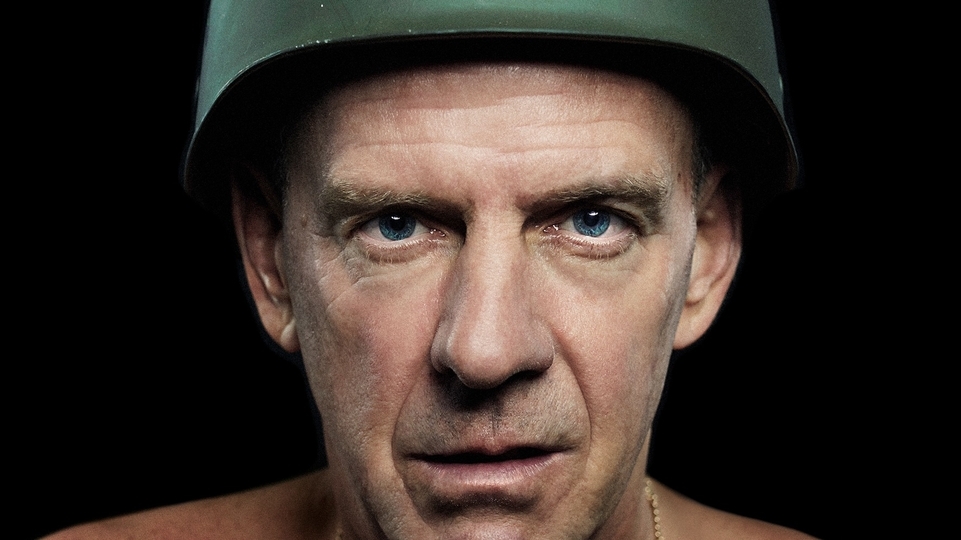 Photo of Fatboy Slim wearing a green army helmet in front of a black background