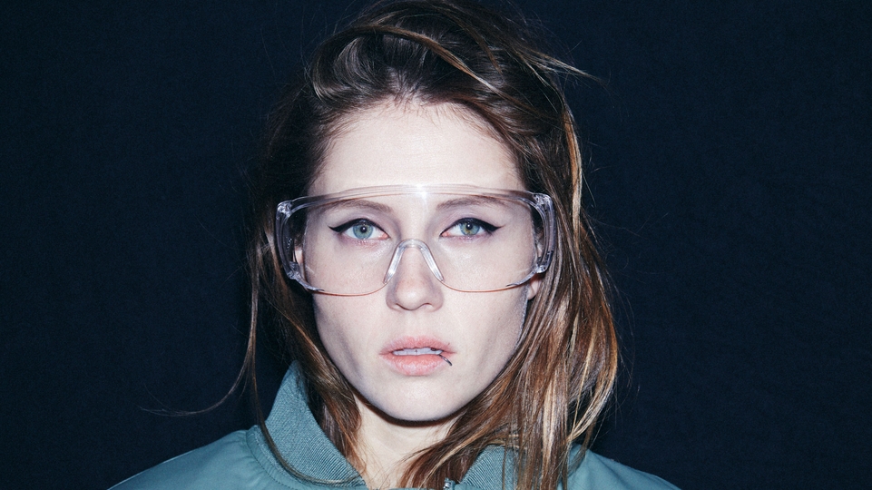 Charlotte De Witte posing against a dark blue backdrop wearing a pair of safety glasses