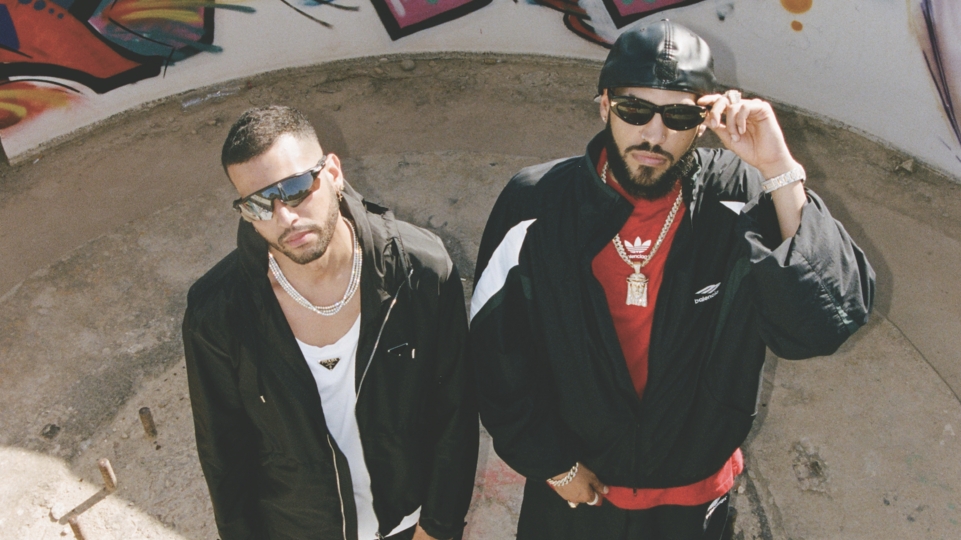 The Martinez Brothers from above standing against a graffiti wall, they are wearing black jackets and sunglasses
