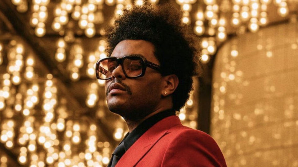 The weeknd poses in a room filled with hanging lights. He's wearing a red suit and large sunglasses