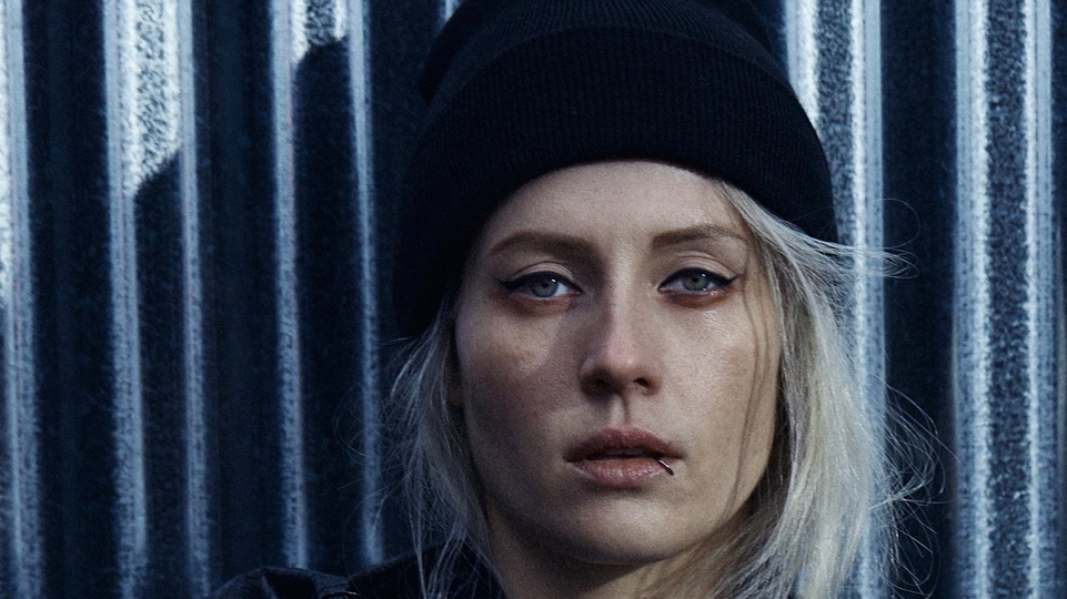 Photo of Charlotte de Witte posing in front of a blue metal wall wearing a black beanie cap