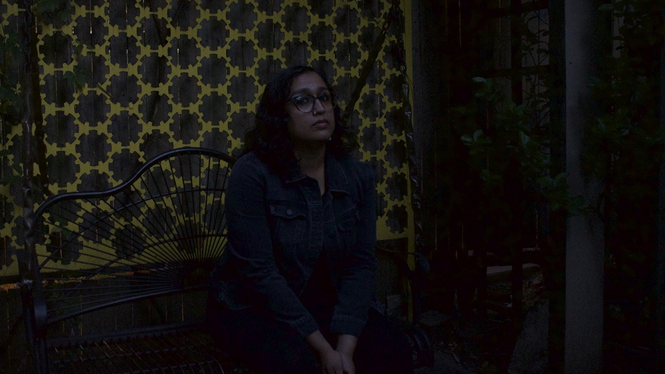 Photo of Krithi sitting on a black bench at night in front of a yellow and brown patterned curtain