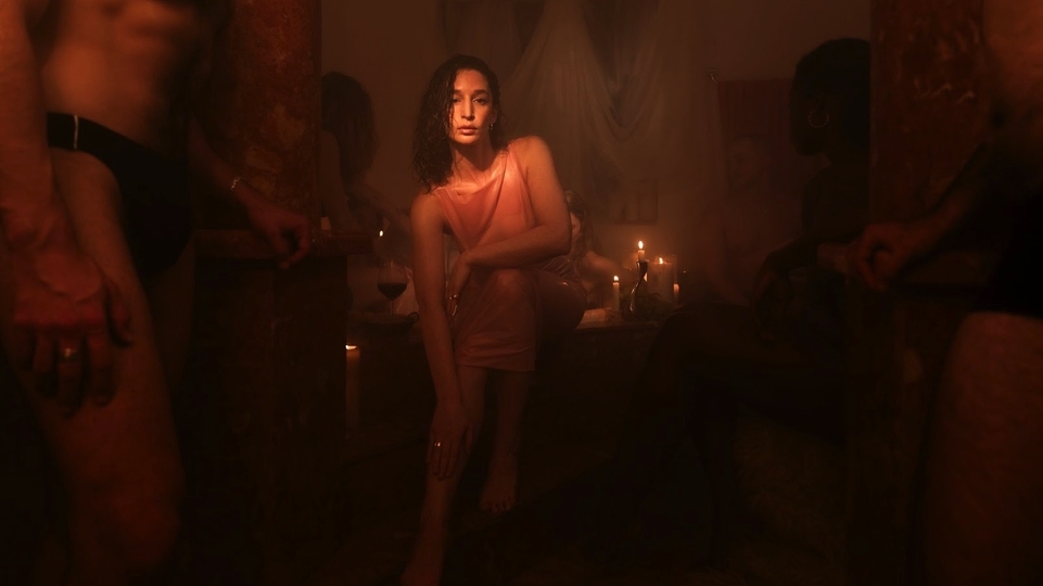 Elkka wearing a pink dress in a steamy room filled with candles. A person in tight black briefs is toward the front of the photo on the left