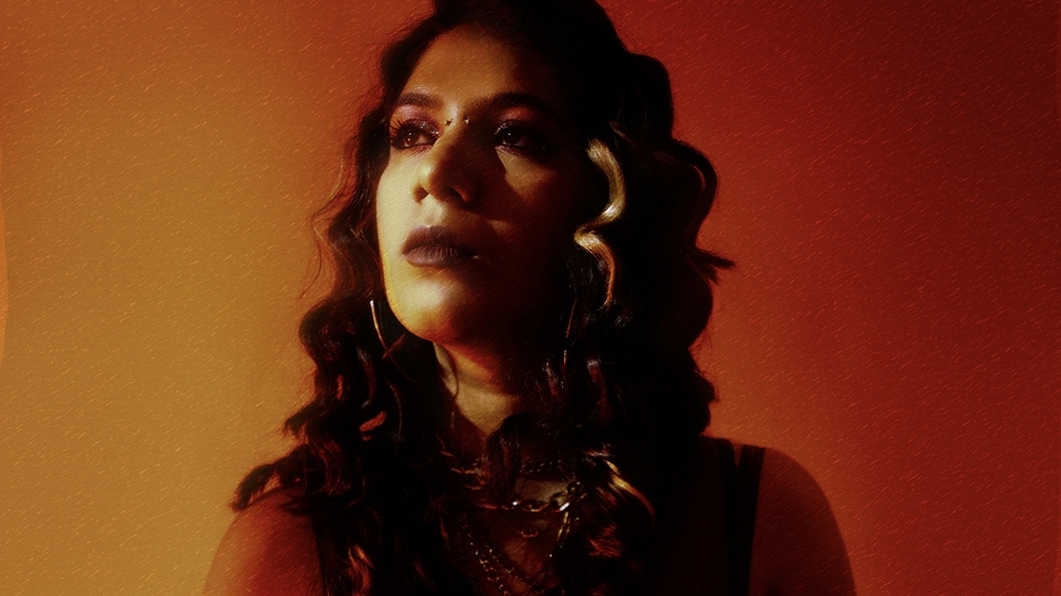 Photo of Zequenx wearing a black dress and heavy jewellery against an orange background 