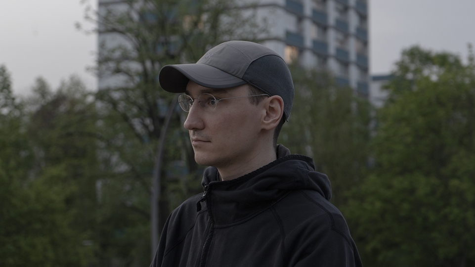 Photo of buttechno standing outside on an overcast day near an apartment block and trees. He's wearing a grey baseball cap and glasses