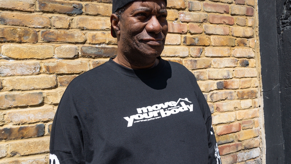 Marshall Jefferson poses in front of a brick wall wearing a black backwards baseball cap and a black long-sleeve t-shirt that says "move your body"